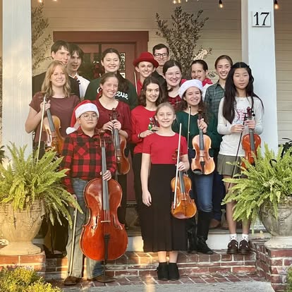 Student musicians in holiday attire posing with violins and cellos on a front porch